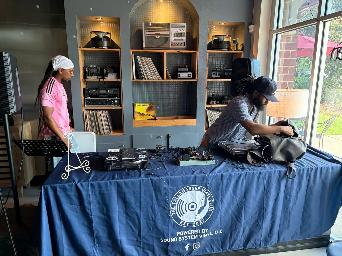 Two DJs at the club table, shelves of records and vintage stereo gear behind them