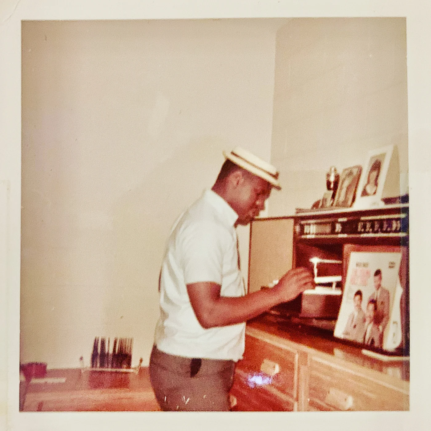 A vintage snapshot of the owner’s father in a straw boater hat, lowering the needle onto a record at a wooden console hi-fi, framed family photographs on top